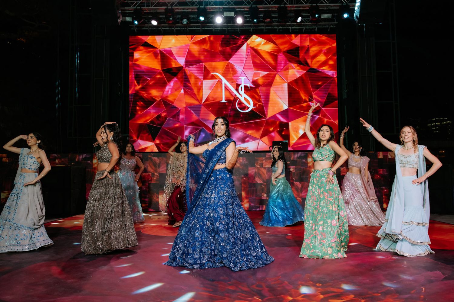 Models showcase vibrant Indian bridal lehengas on a runway with a red geometric NS logo backdrop at a fashion show.