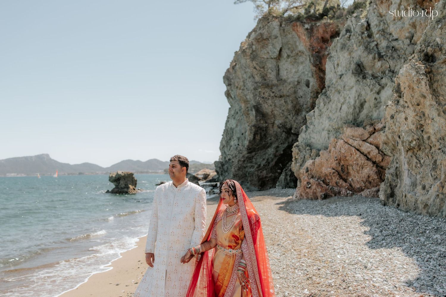 Indian bride in red lehenga and groom in white sherwani walk hand-in-hand on a scenic rocky beach.