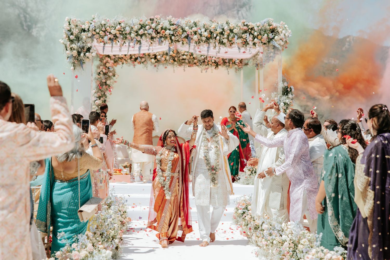 Bride and groom exit under floral mandap at vibrant Indian wedding ceremony with guests celebrating.