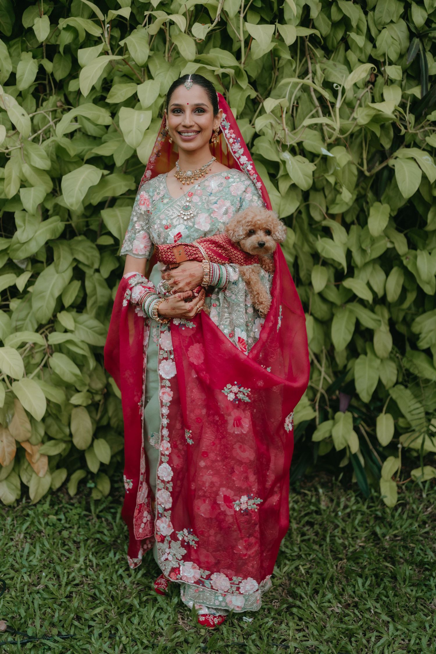 Bride in mint green and red floral lehenga holding a small dog, standing before lush green foliage backdrop.