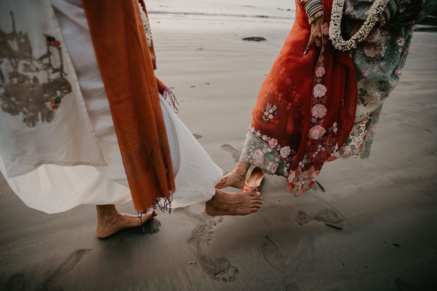 Two women in vibrant traditional Indian attire walk barefoot on sandy beach, showcasing intricate henna and wooden sandals.
