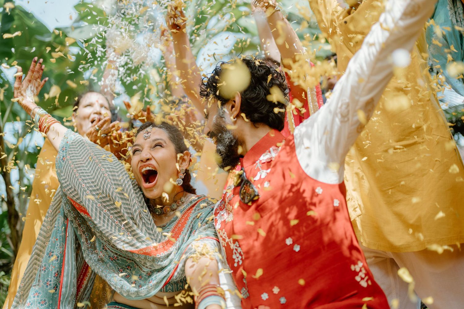 People in colorful traditional attire celebrate joyfully with golden confetti raining down at a festive Indian wedding.