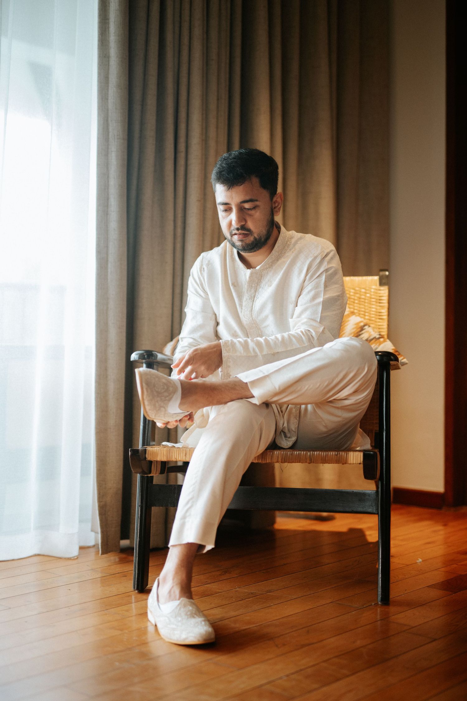 Man in white linen outfit sitting in wooden chair adjusting cufflinks by window with curtains.
