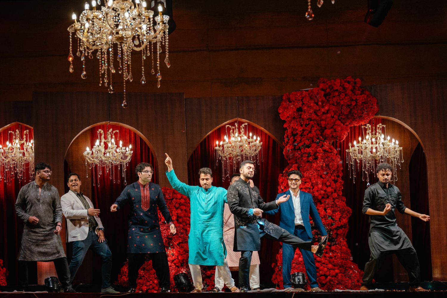 Performers dancing on stage with red floral backdrop and chandeliers at an elegant Indian cultural event.