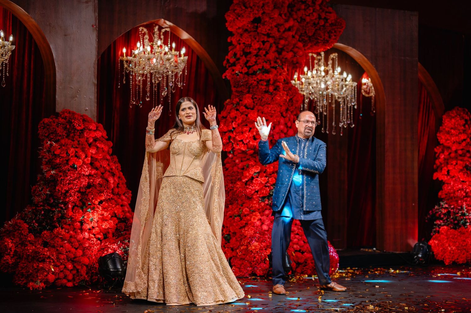 Bride in golden lehenga and man in blue suit wave on stage with red floral backdrop and chandeliers at wedding.