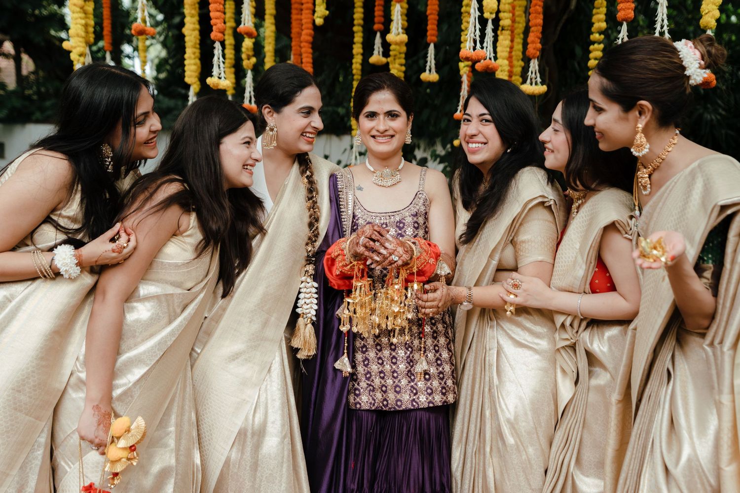Bride in purple lehenga surrounded by bridesmaids in gold sarees, laughing and holding floral bouquet at Indian wedding.