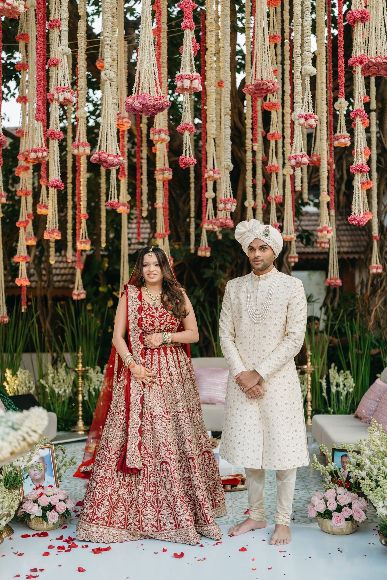 Indian bride in red lehenga and groom in cream sherwani stand under floral decorations at traditional wedding ceremony.
