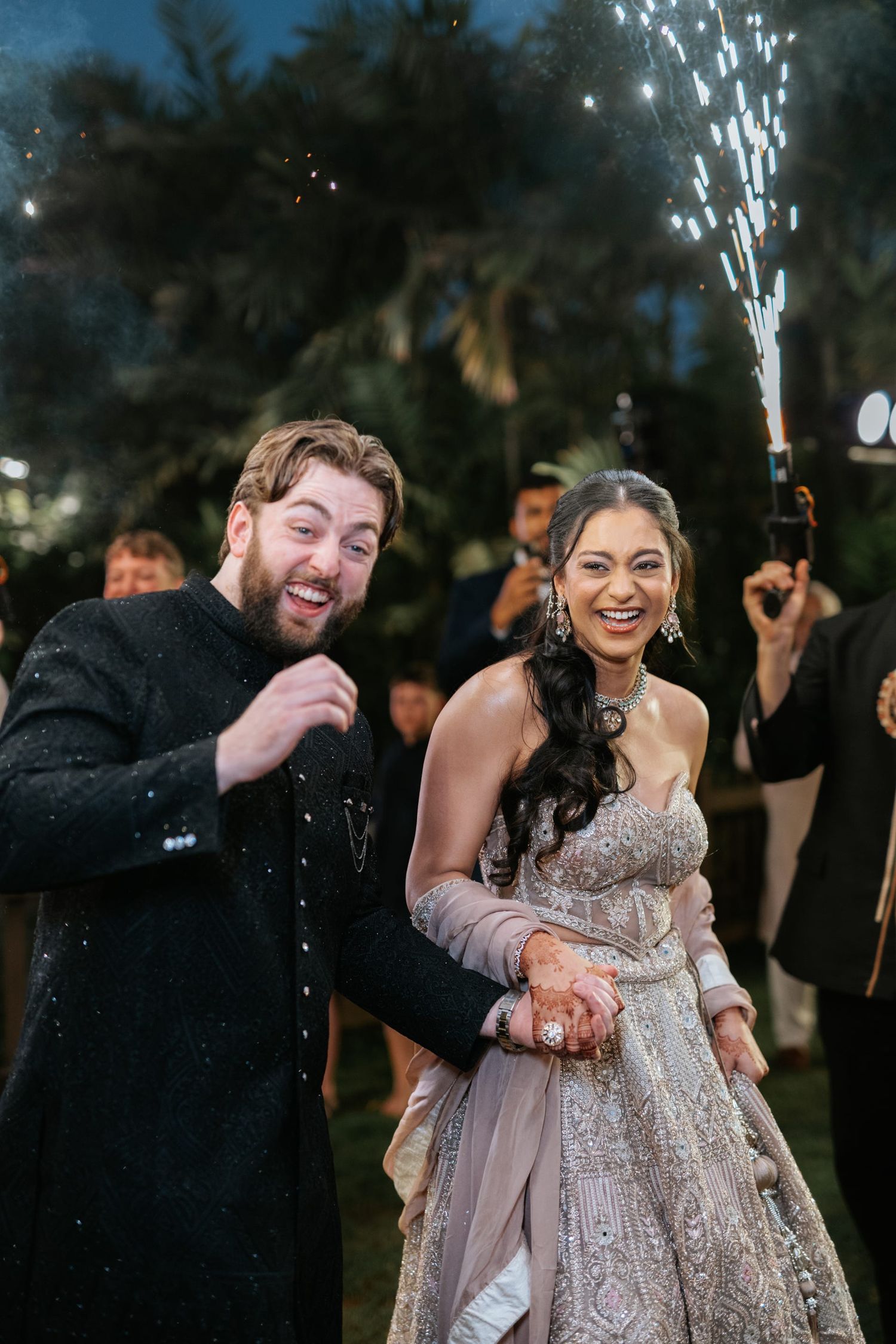 Joyful couple in Indian wedding attire celebrating with sparklers at night outdoor ceremony.