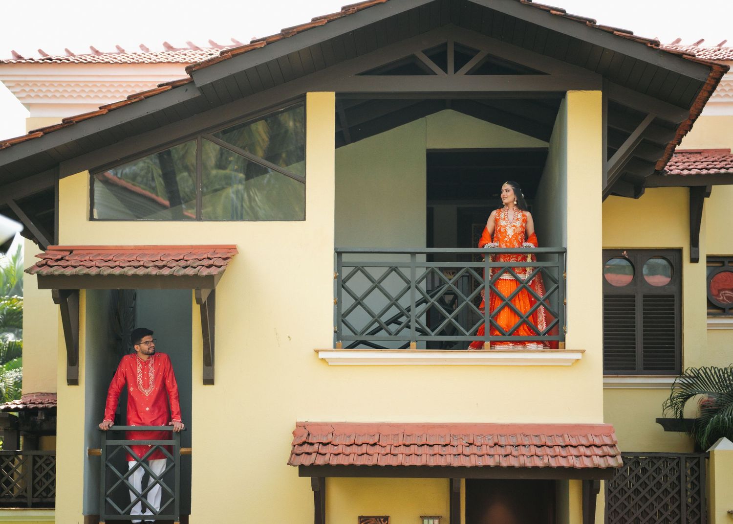 Yellow stucco house with terracotta roof tiles features two people standing on different levels of a covered balcony.