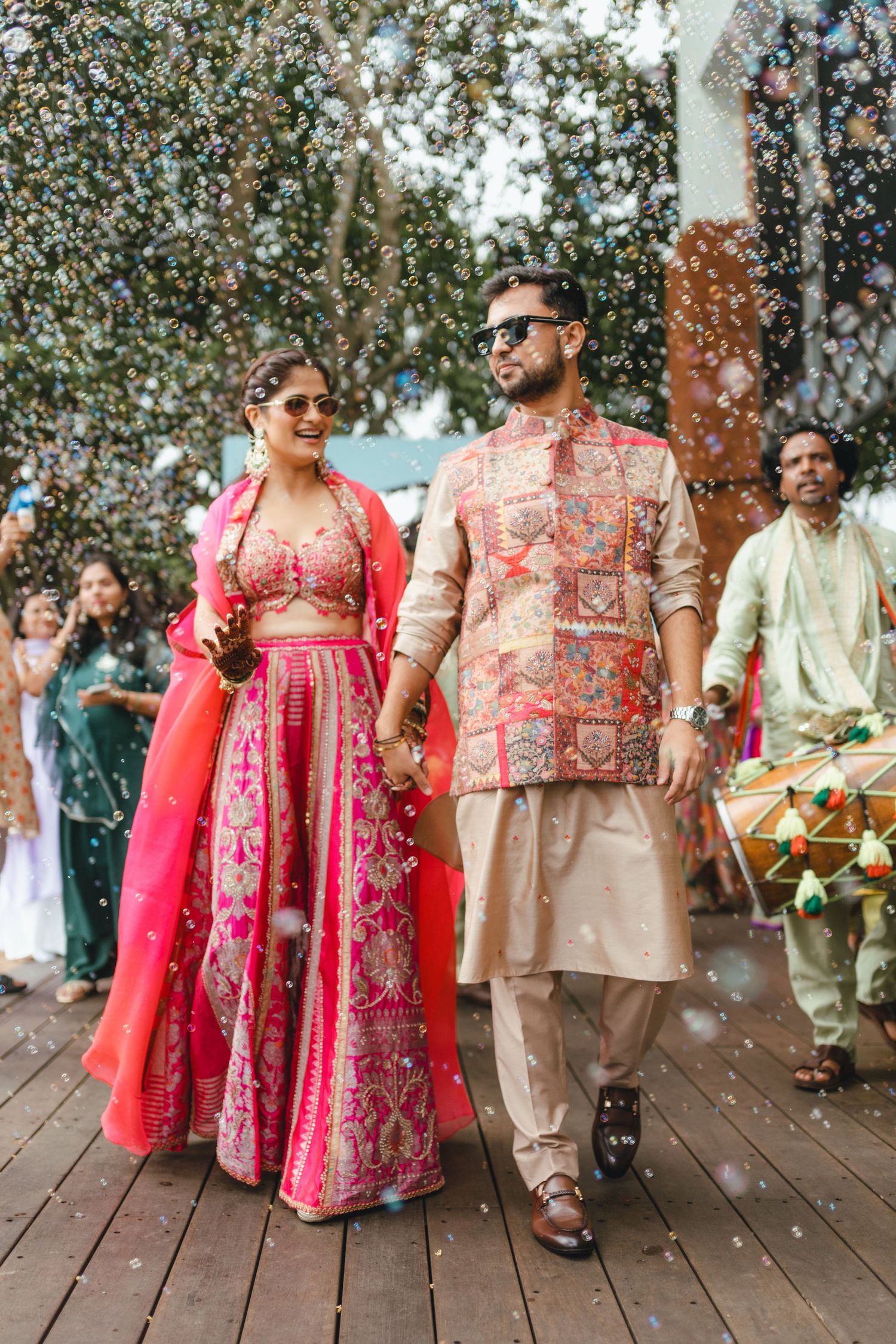 Couple in vibrant Indian wedding attire walk hand-in-hand, woman in pink lehenga, man in gold sherwani, surrounded by bubbles.