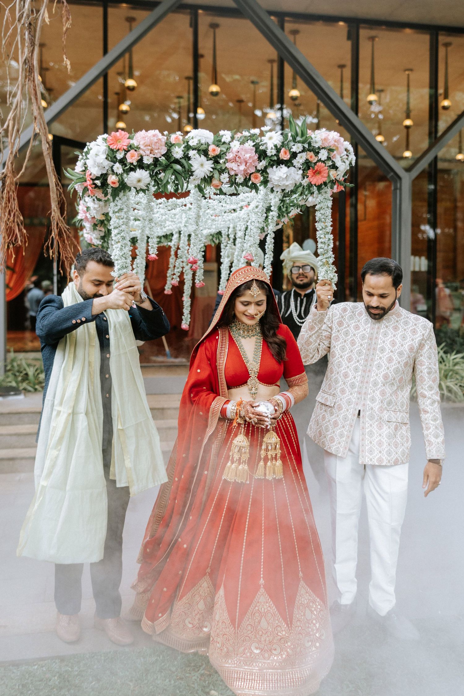Bride in red lehenga walks under floral canopy with family escorts at Indian wedding ceremony.