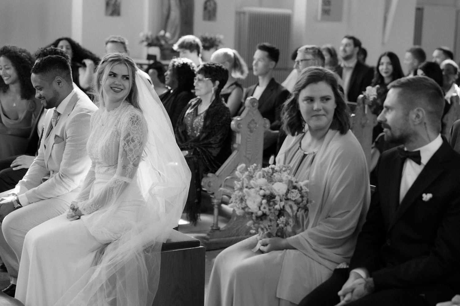 Wedding party and guests seated in formal attire during ceremony in black and white.