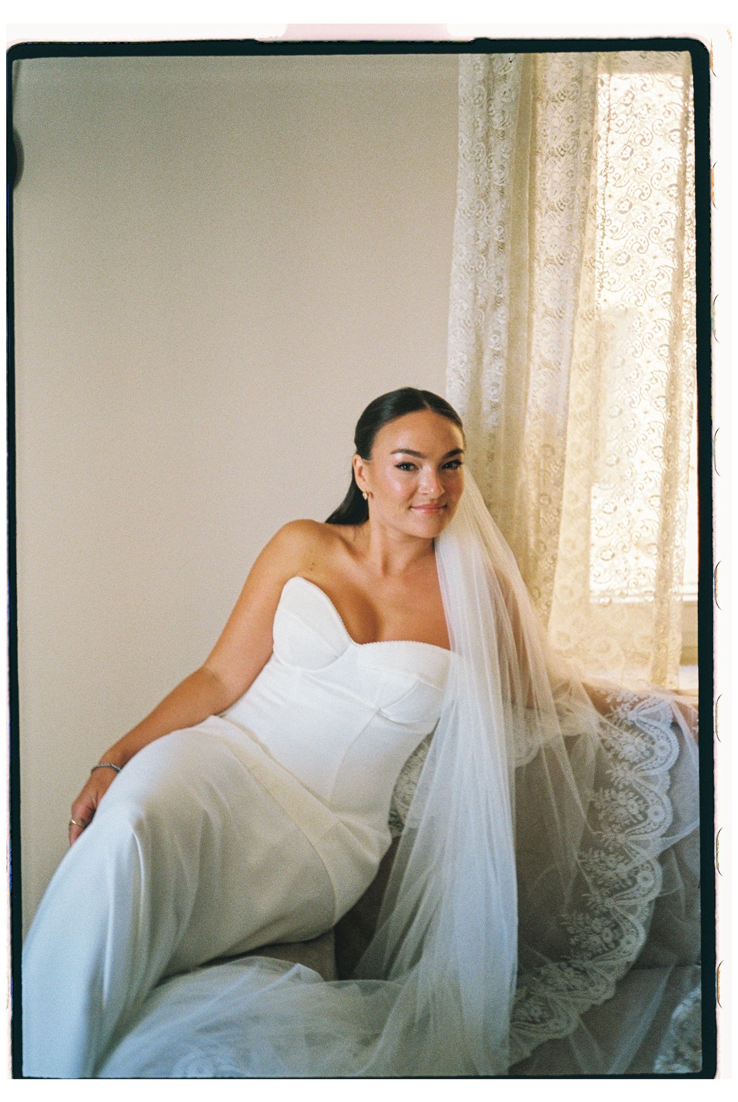 Bride in strapless white gown and lace veil sits by lace curtained window.