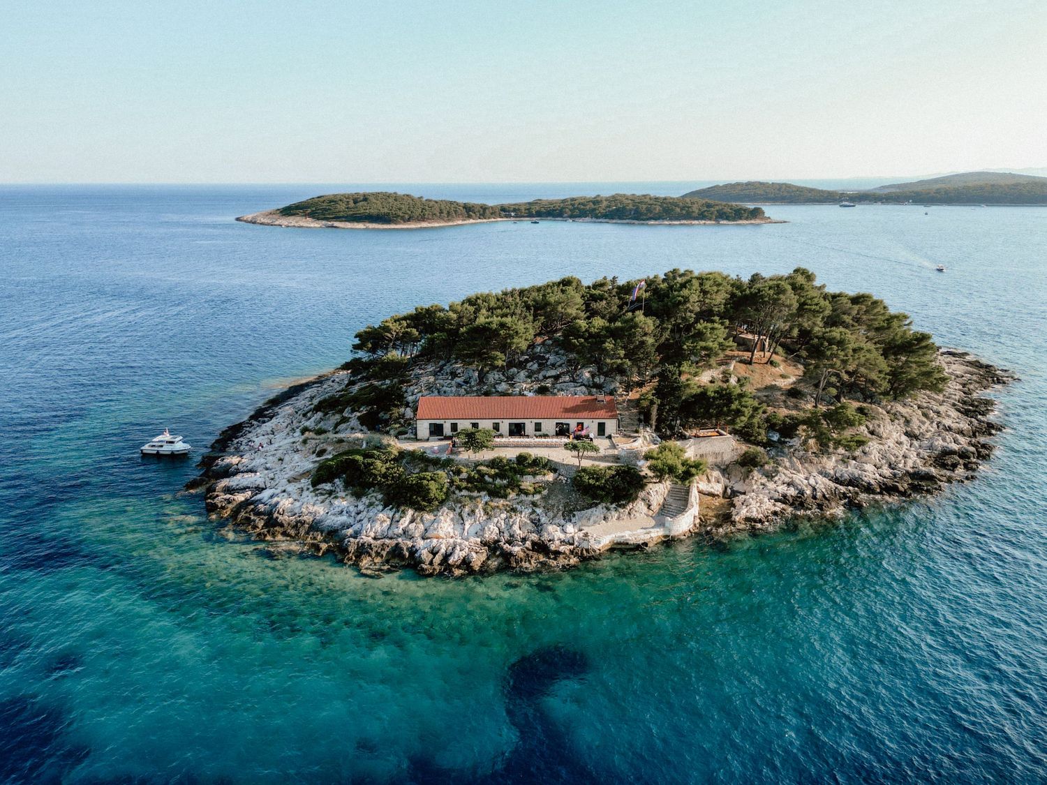 Aerial view of a small rocky island with red-roofed villa surrounded by turquoise Adriatic Sea waters near Croatia.