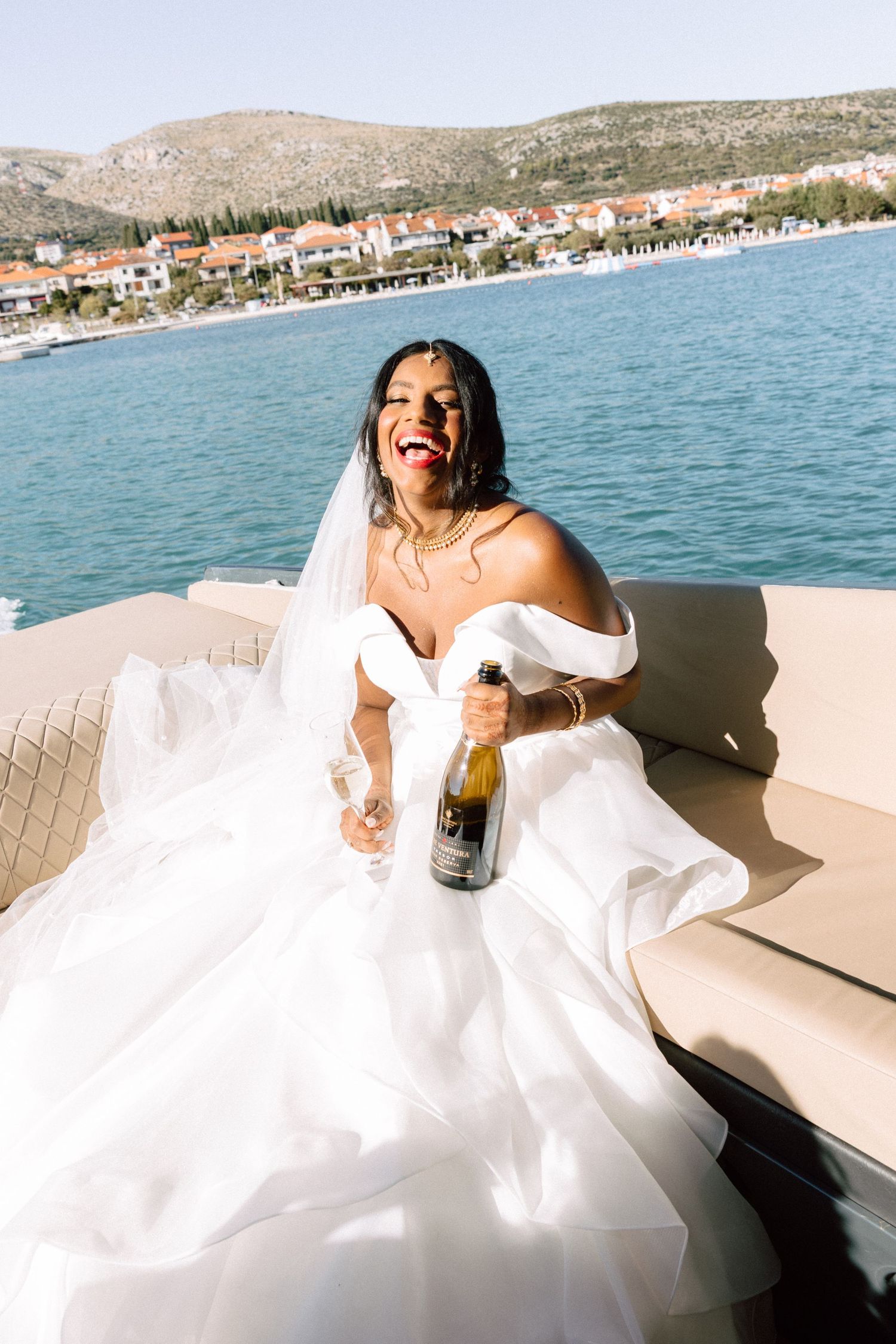 A joyful bride in an off-shoulder white gown laughs while holding a champagne bottle on a scenic waterfront boat.