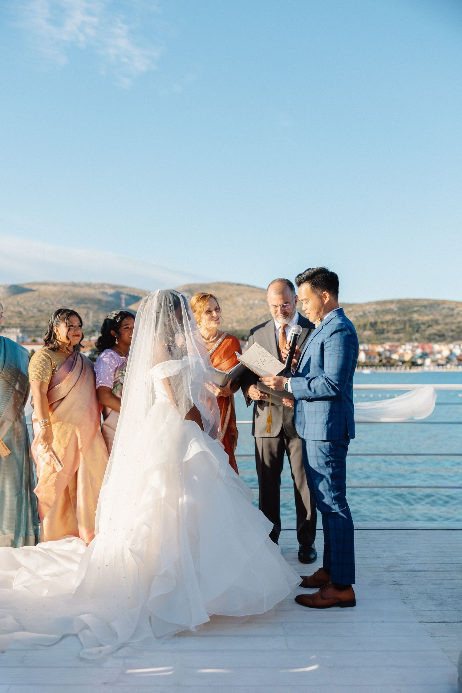 Bride and groom exchanging vows on a waterfront pier with bridesmaids and officiant under blue sky.