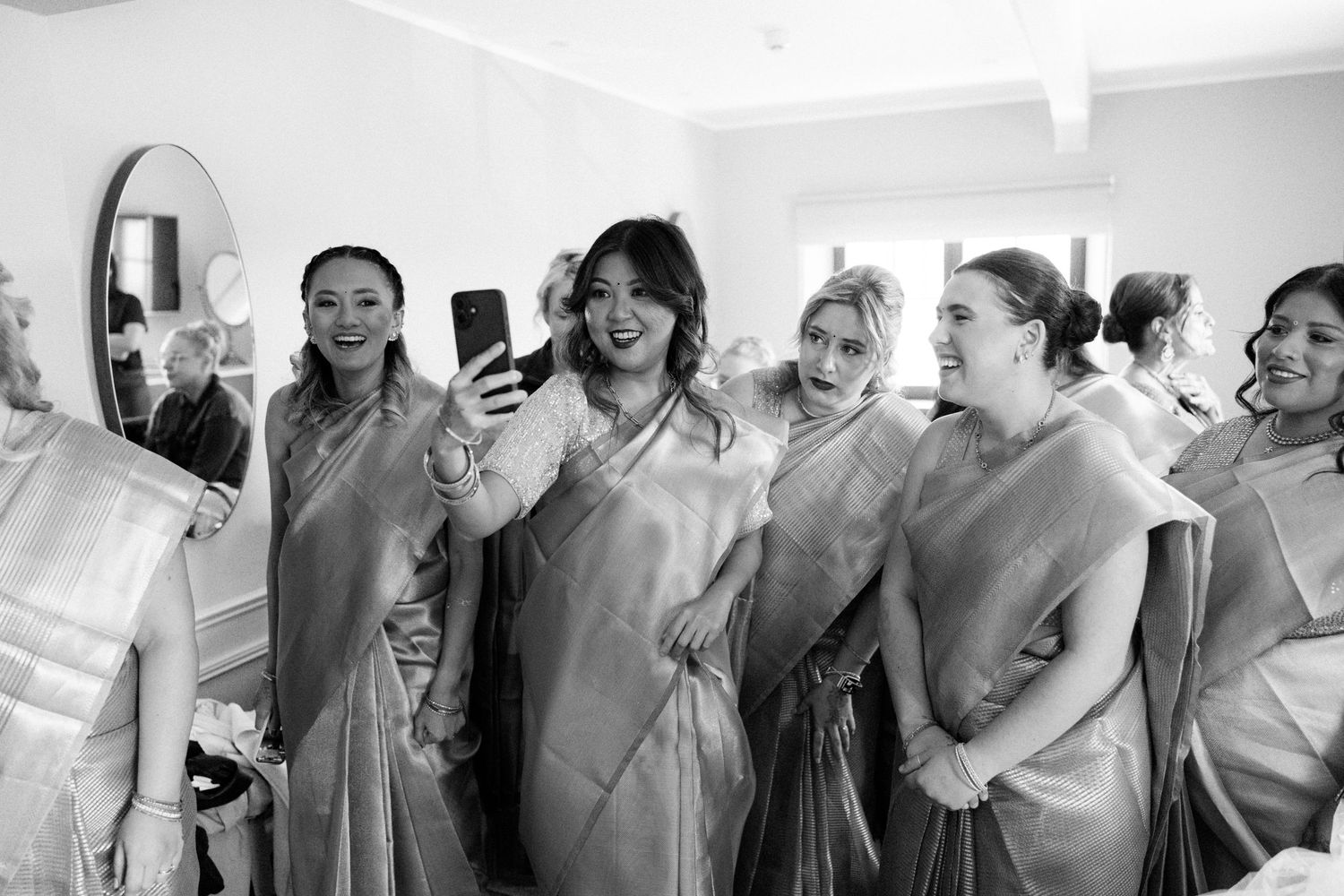 Bridesmaids in matching sarees laughing and taking a selfie together while getting ready for a wedding.