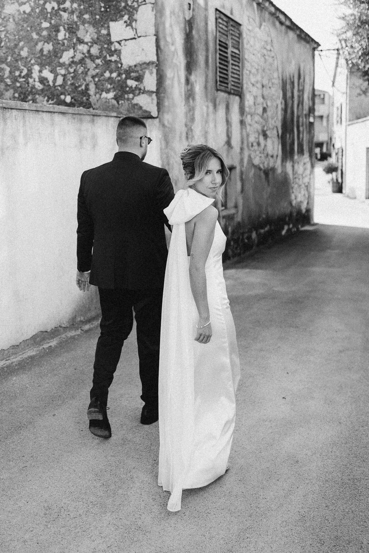 A bride in a sleek white gown glances back while walking down a rustic alley with textured walls.