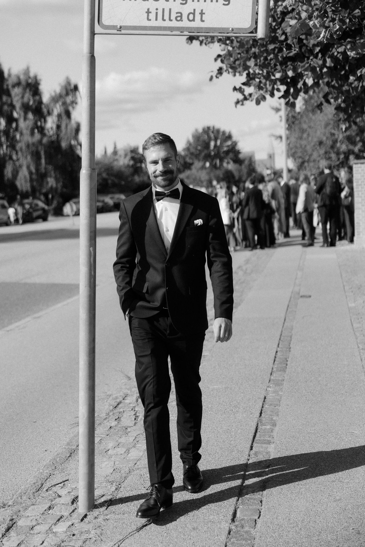 Black and white photo of person in tuxedo walking along street with signpost.