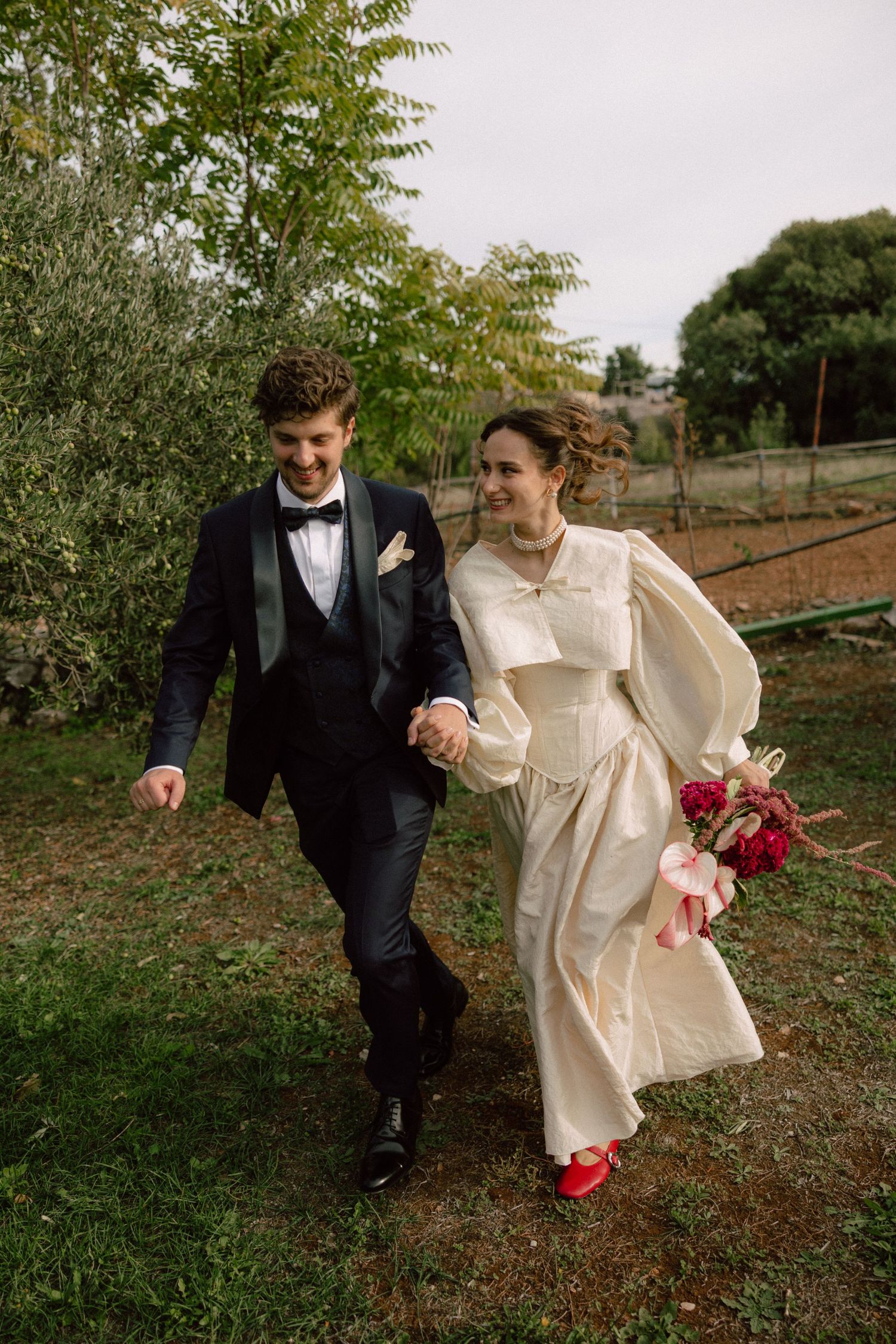 A couple in wedding attire walks together outdoors with the bride wearing a cream coat and red shoes.