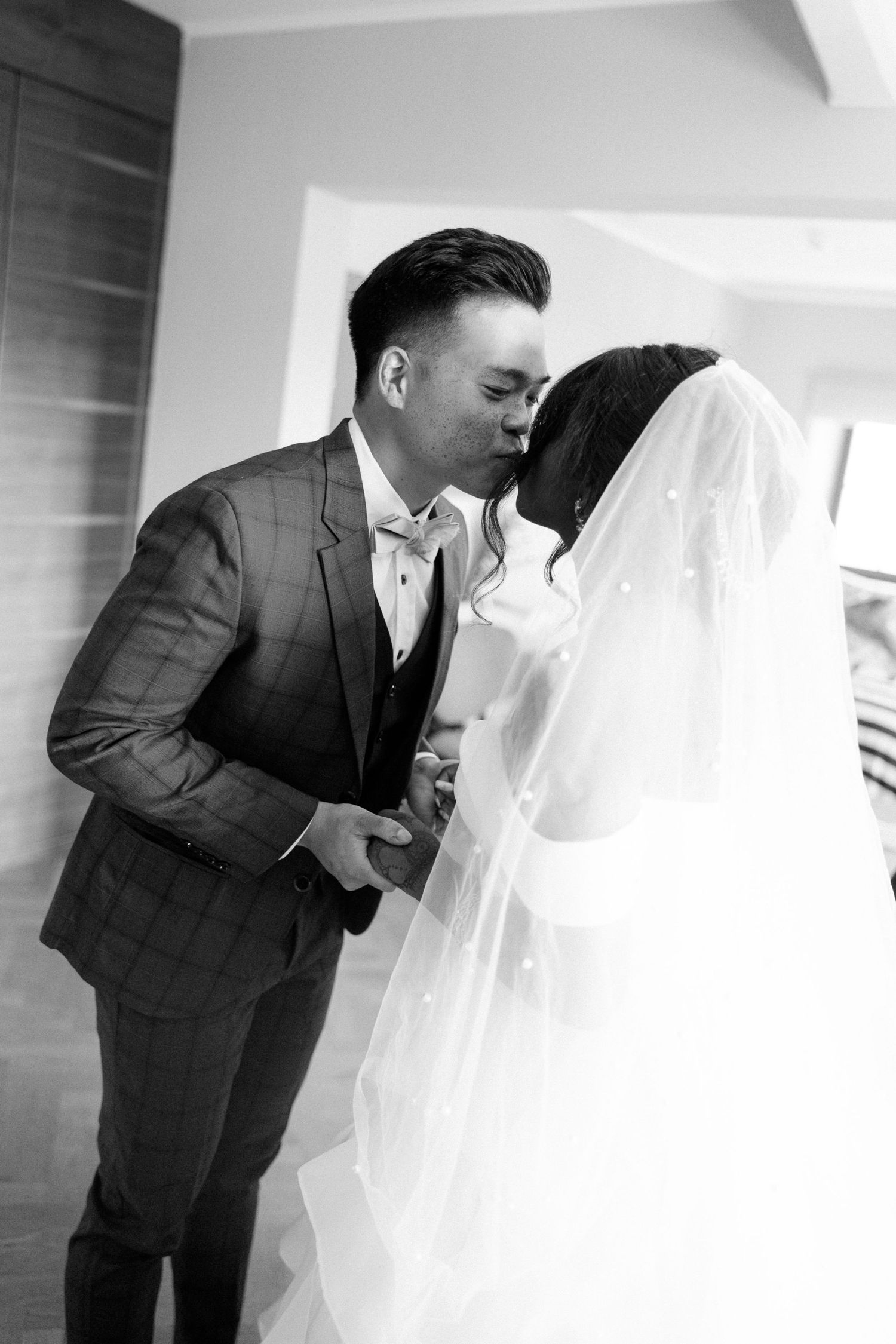 Black and white photo of a groom in a plaid suit kissing his veiled bride indoors.