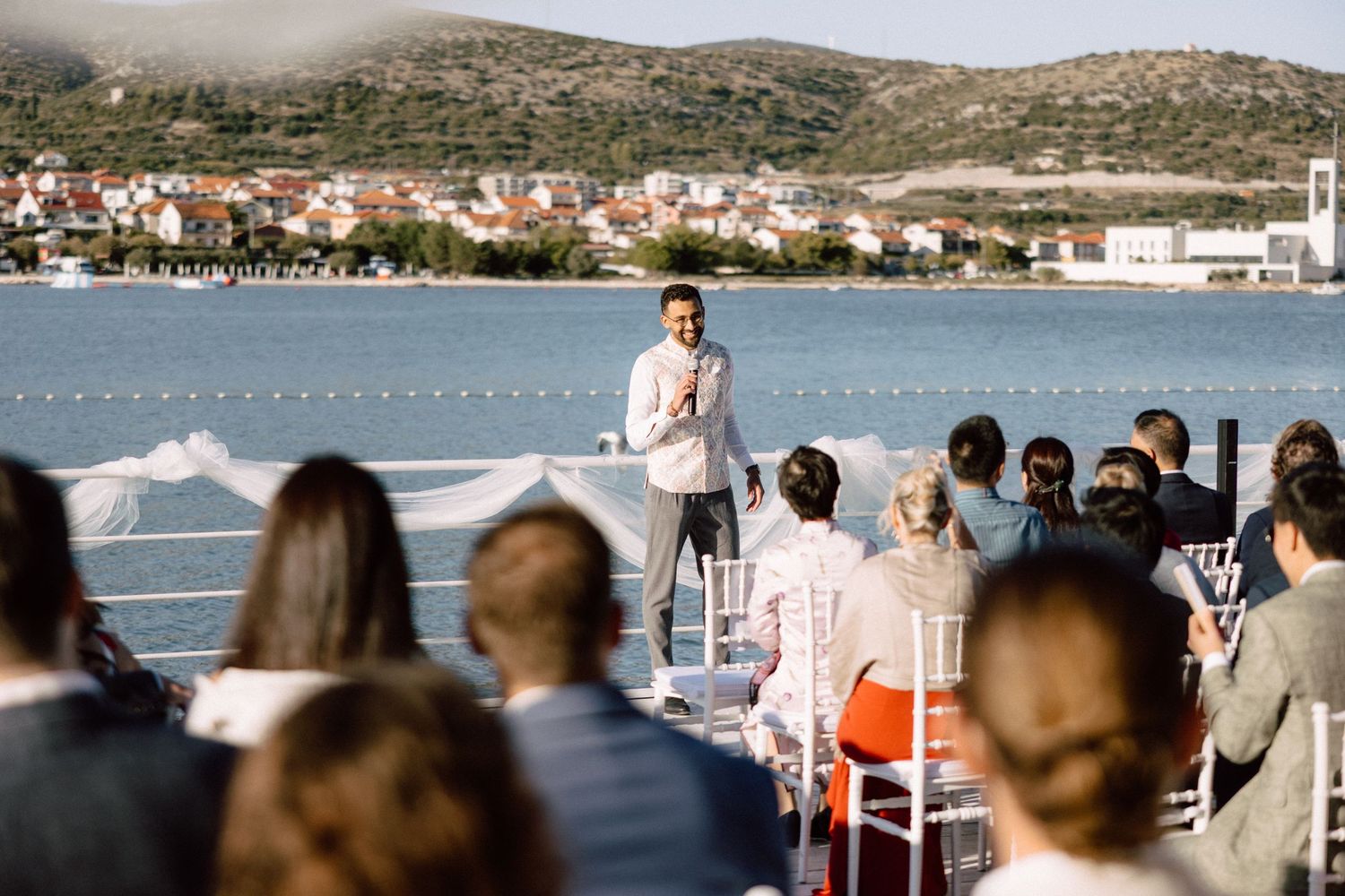 Groom stands at an outdoor waterfront wedding ceremony altar addressing seated guests.