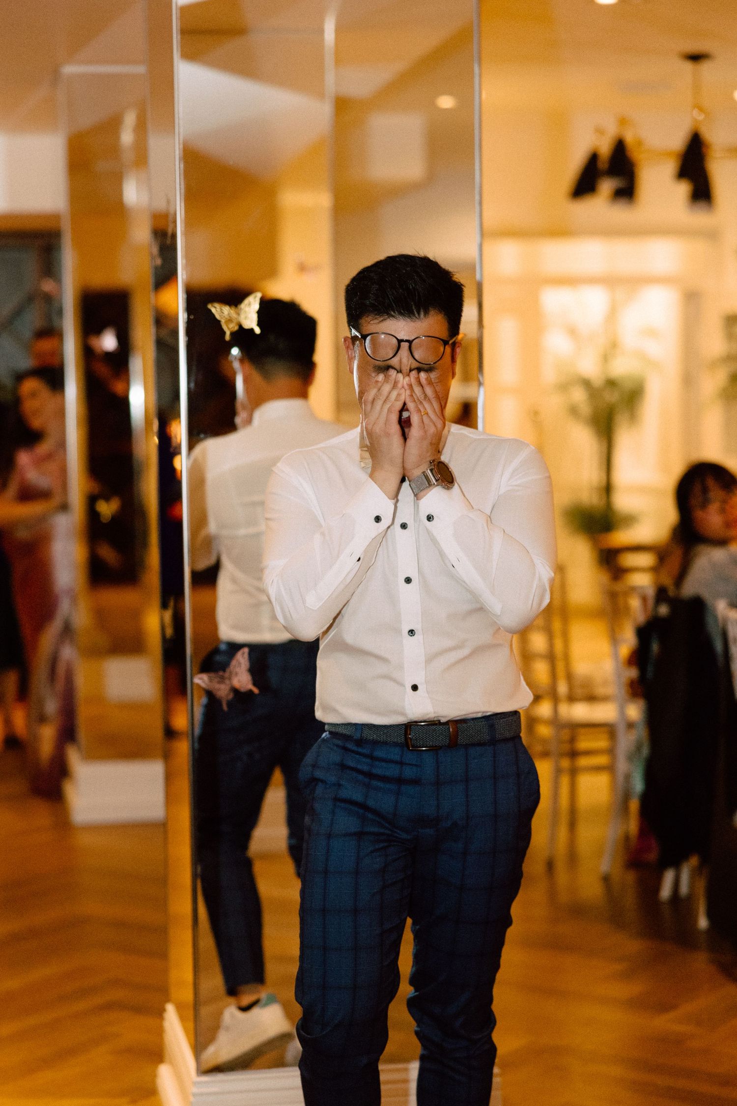 Emotional man in glasses and white shirt covering his mouth with hands in surprise at a wedding event.