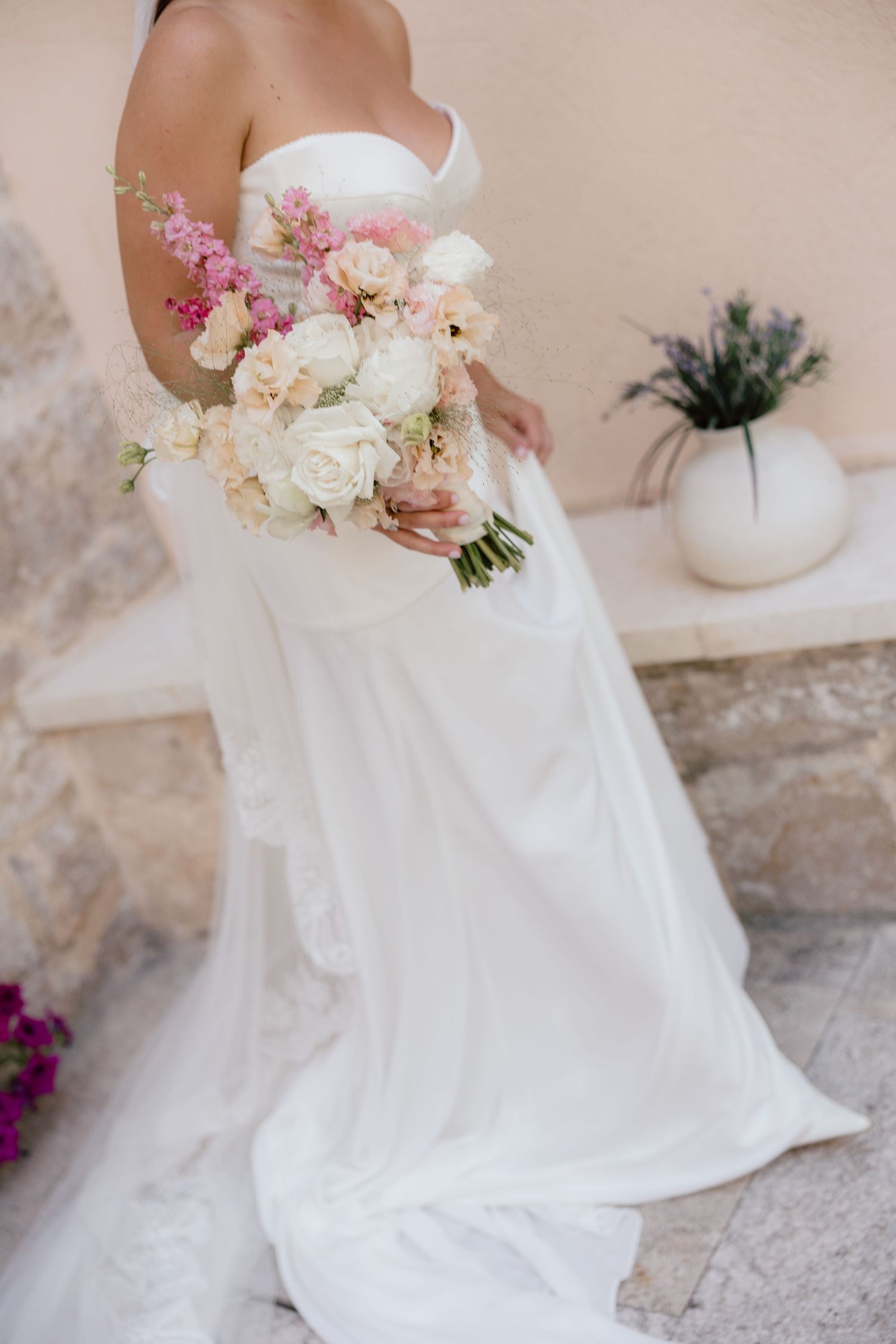 Bride in white strapless wedding gown holding a bouquet of white and pink roses at an outdoor venue.