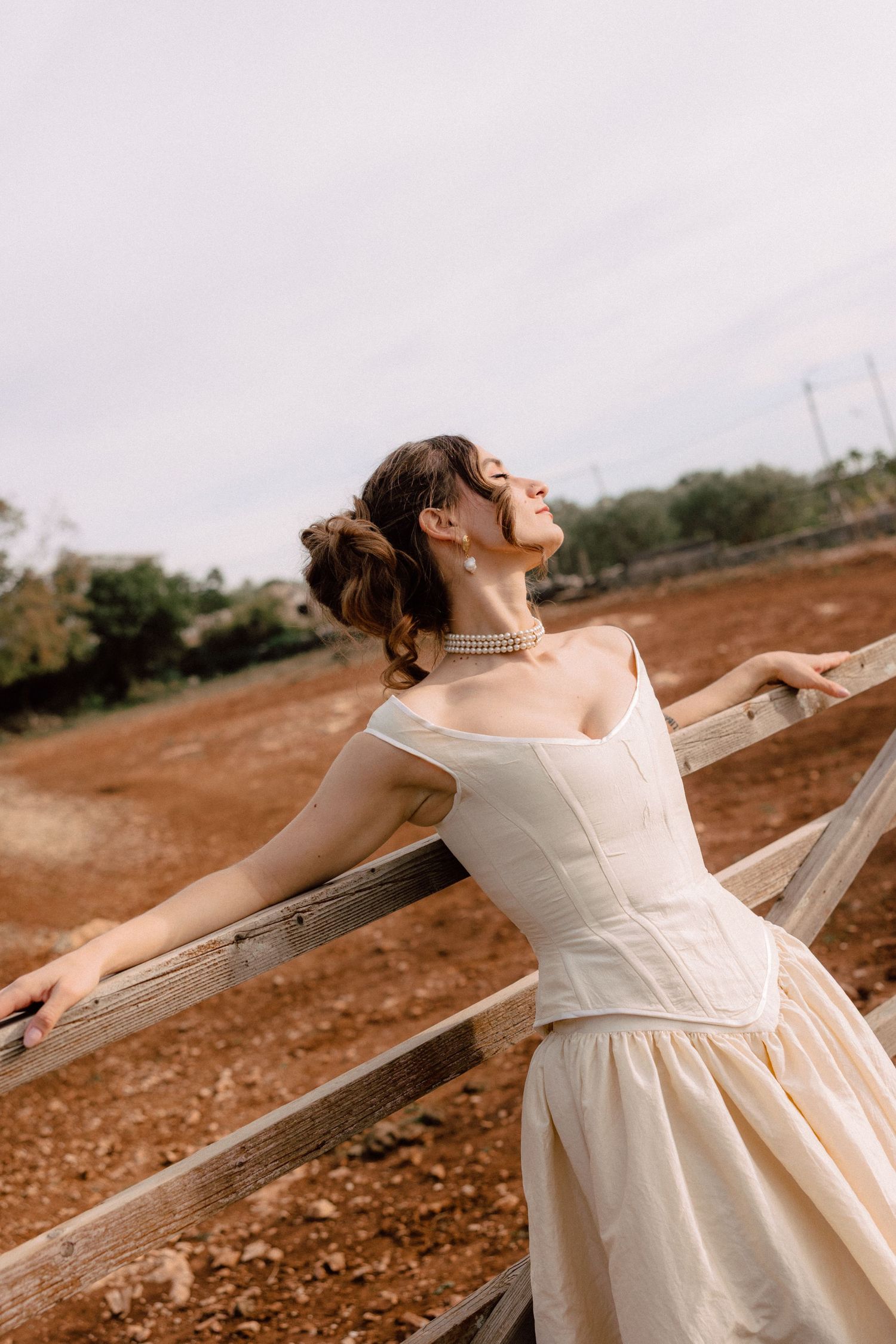 Bride in vintage-style dress poses joyfully against rustic outdoor backdrop.