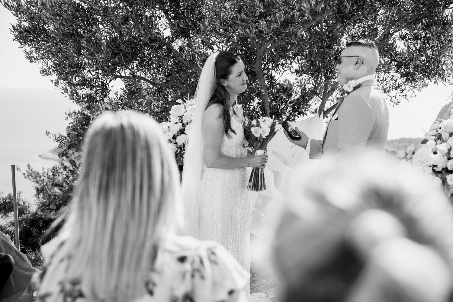 Wedding guests seated at outdoor ceremony under trees with soft natural lighting.