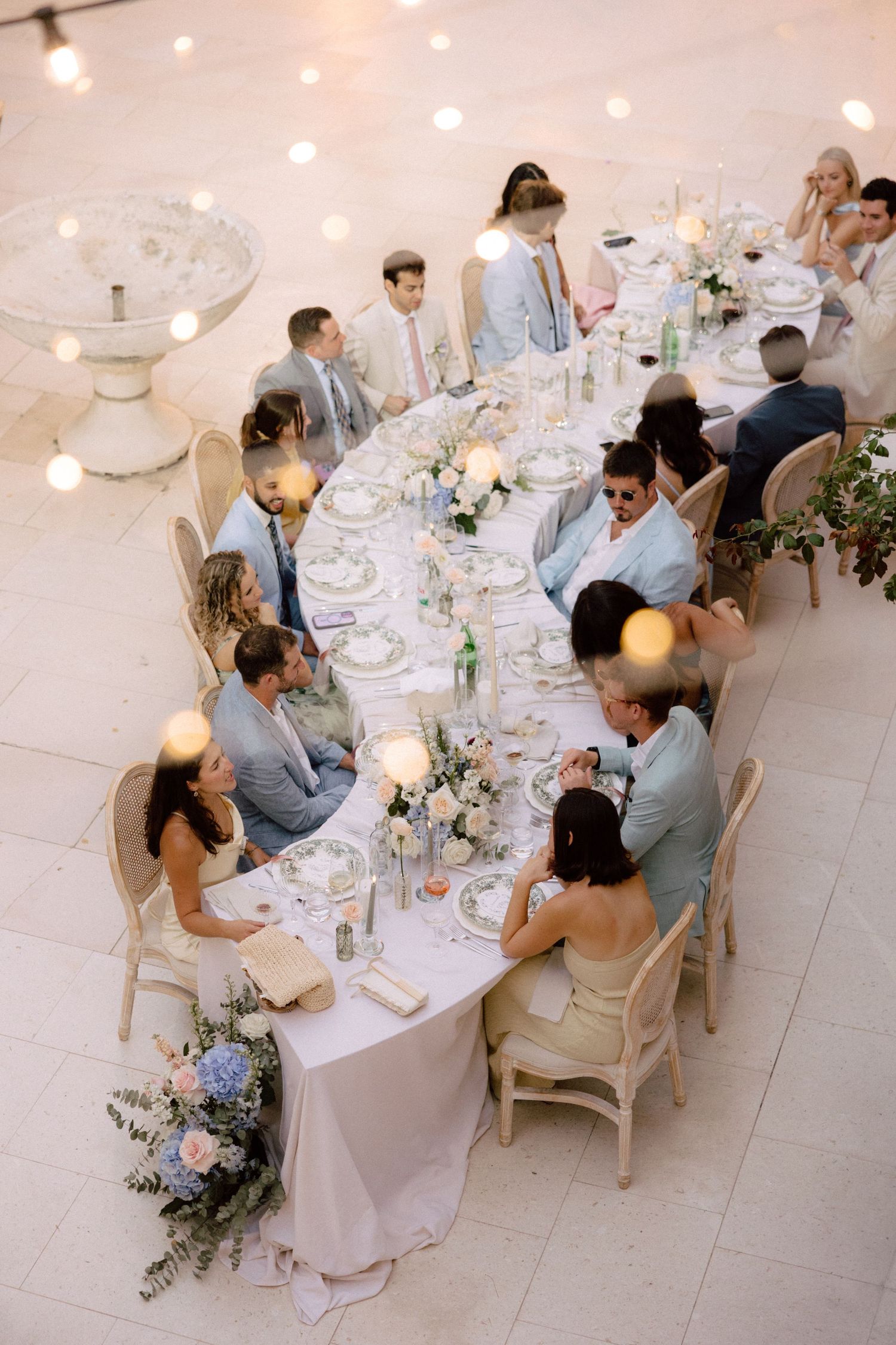 Elegant wedding reception dinner table with white linens, floral centerpieces and string lights creating an intimate atmosphere.