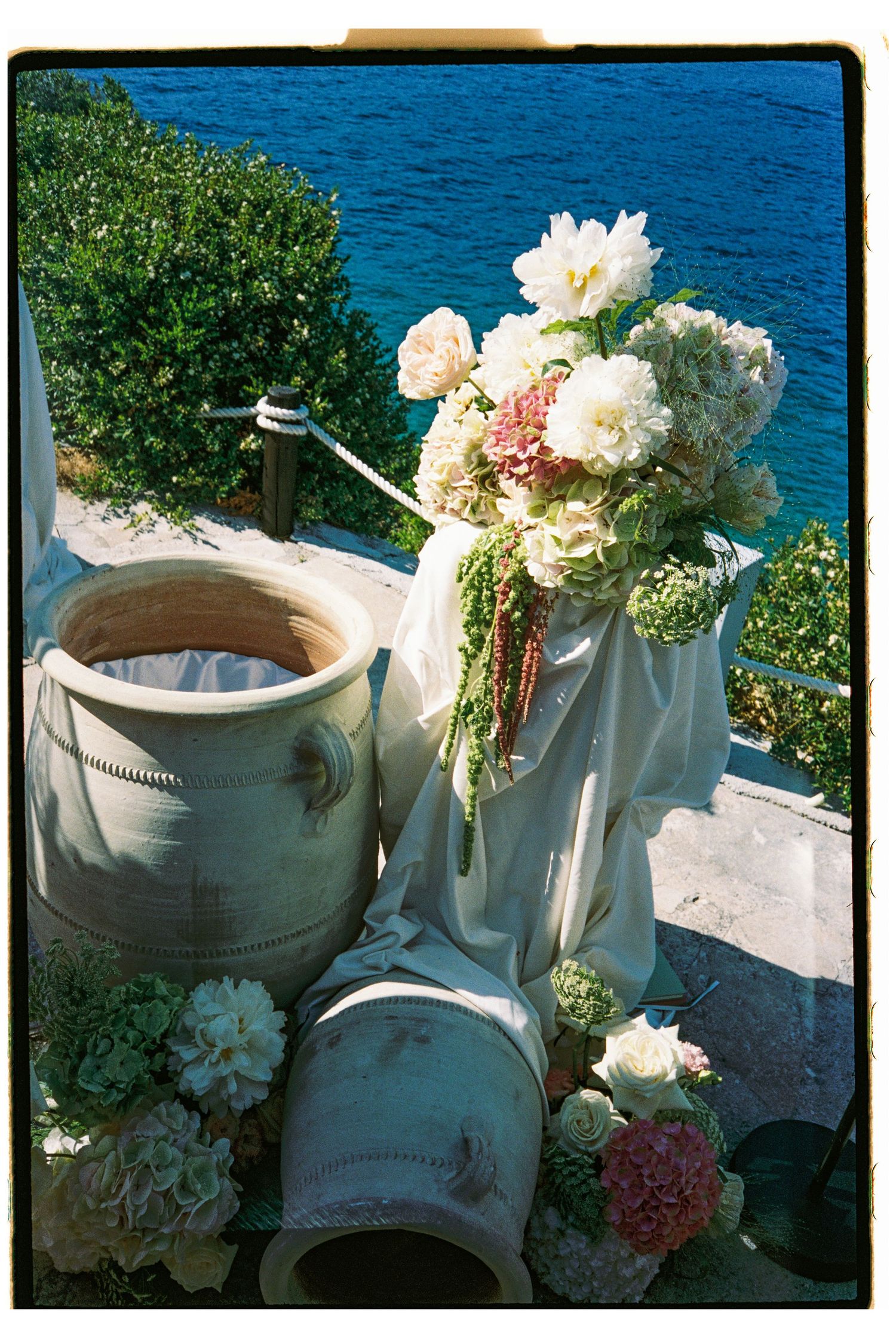Elegant wedding floral arrangement with white peonies and pink hydrangeas beside large ceramic urns overlooking turquoise sea.