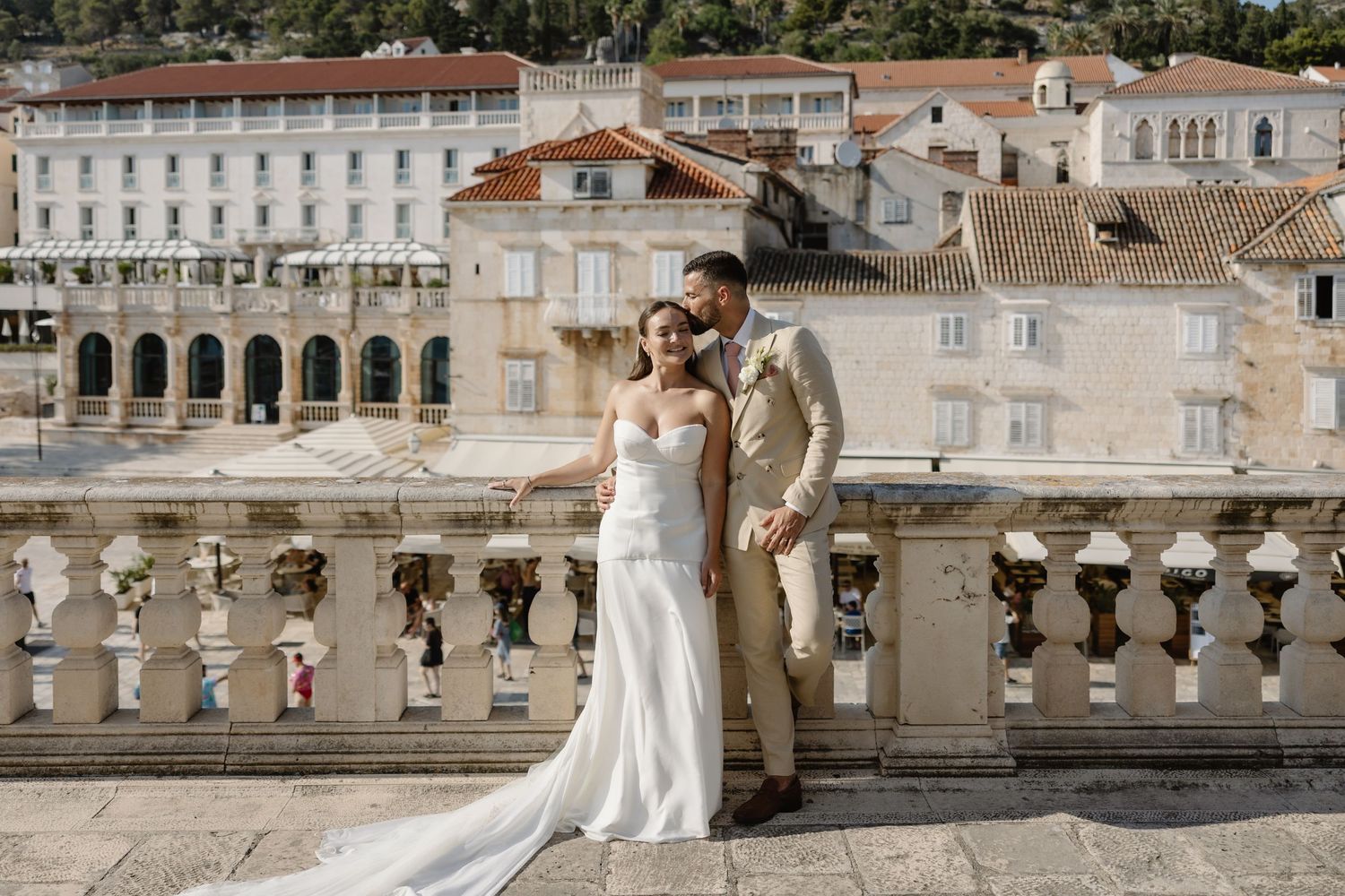 Bride and groom pose on a stone balcony overlooking historic European architecture on their wedding day.