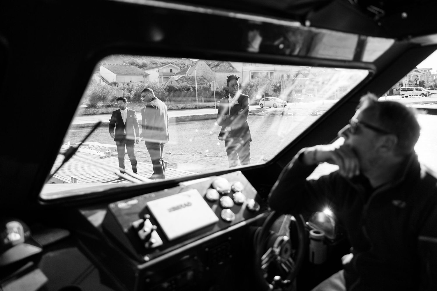 Black and white photo of boat captain steering vessel with wedding guests visible through window.