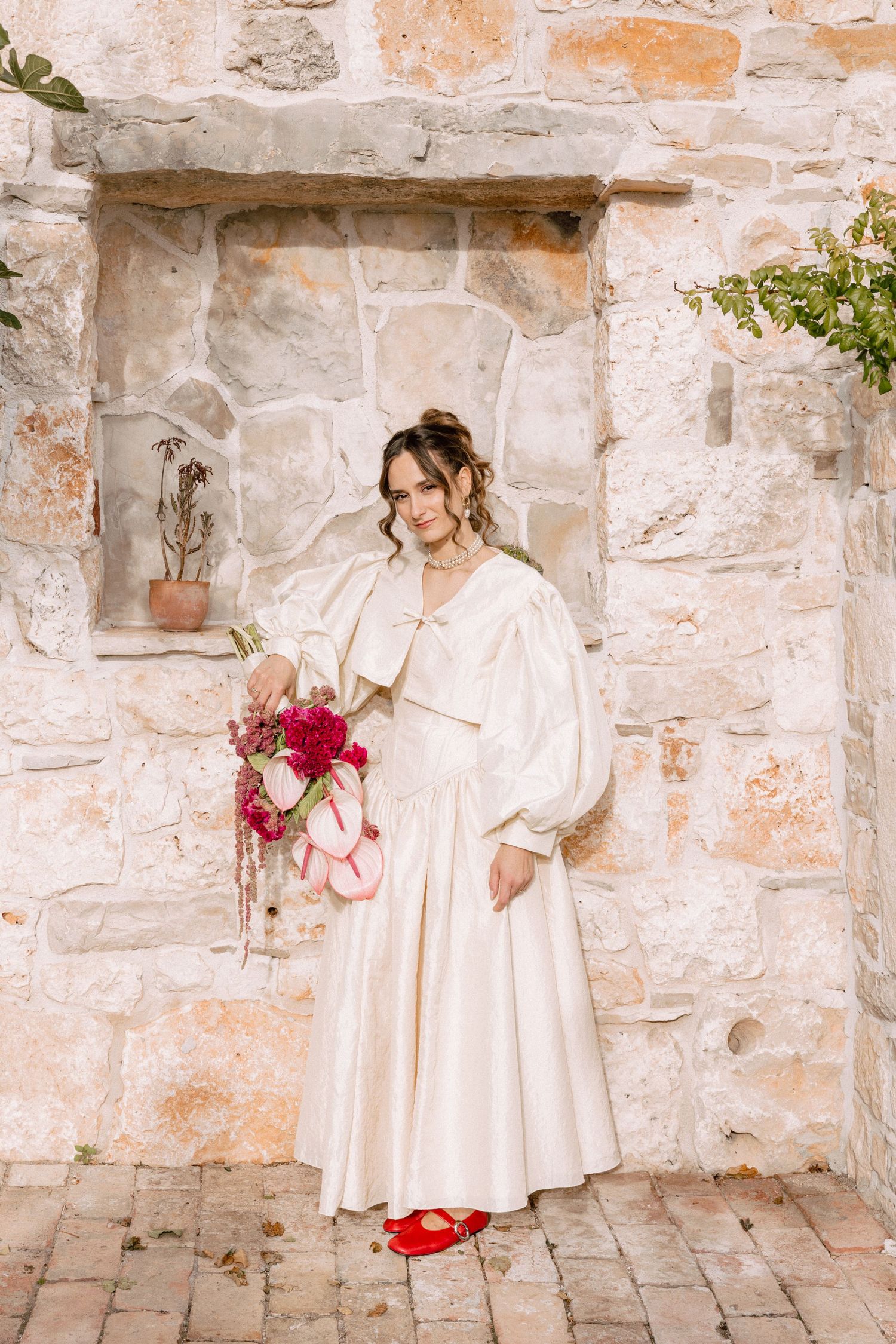 Young children in cream-colored dresses play and interact along a rustic stone wall with pink floral accents.