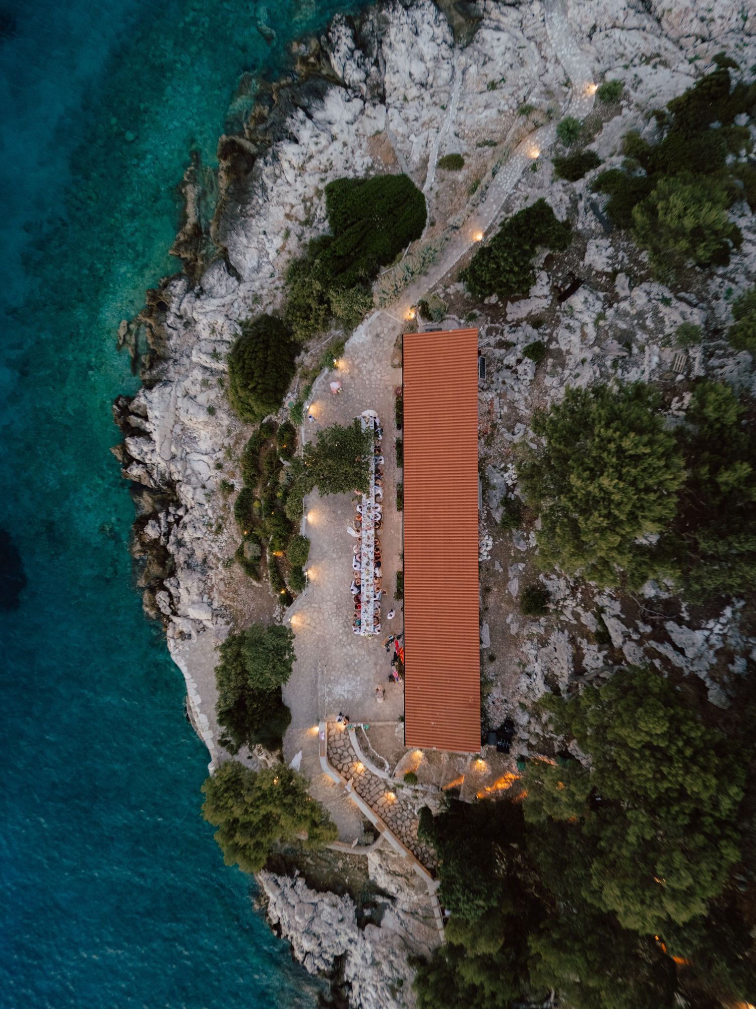 Bird's eye aerial view of illuminated wedding venue with terracotta roof on rocky coastline at twilight.