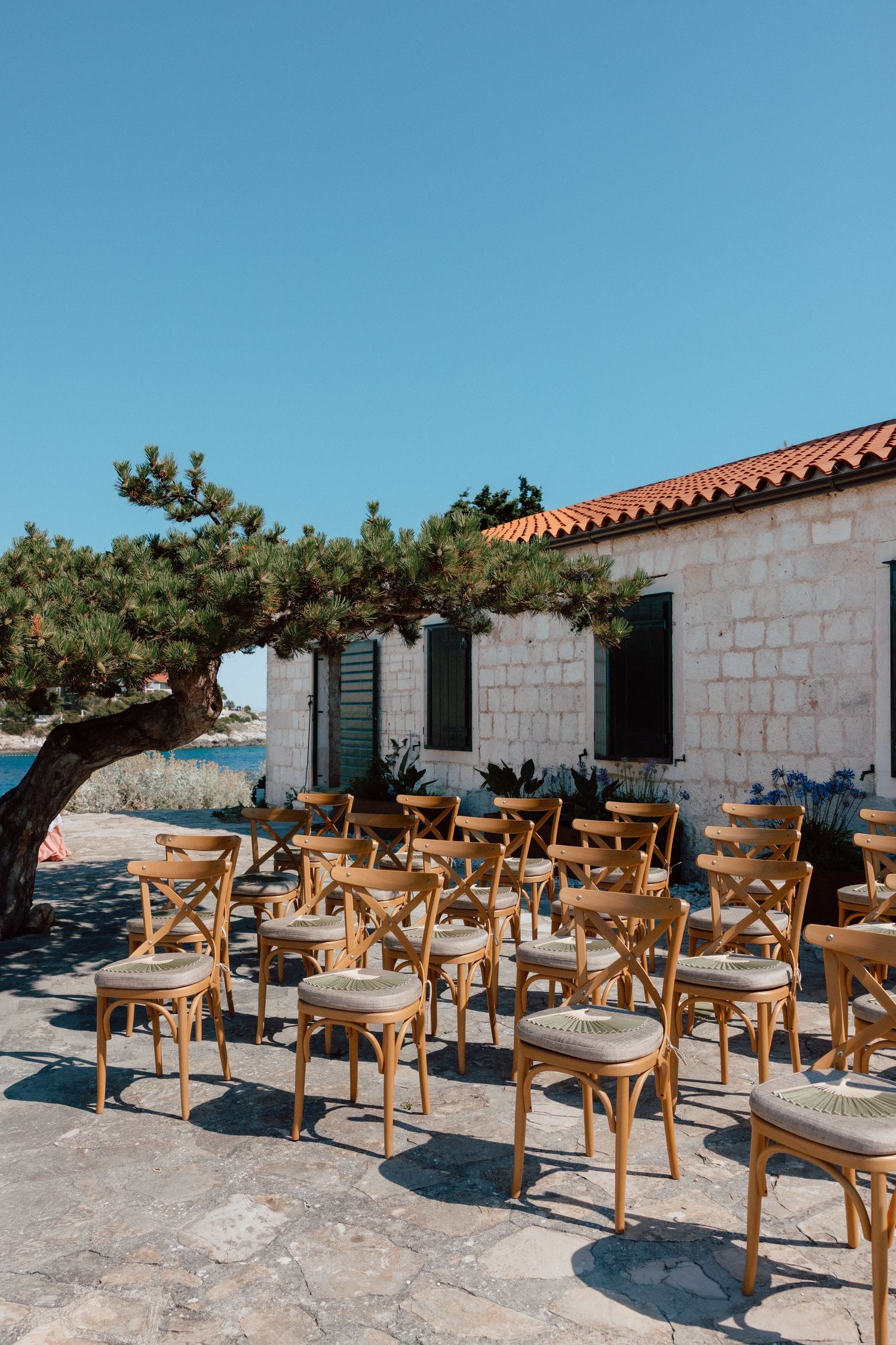 Outdoor wedding ceremony setup with wooden cross-back chairs on stone terrace overlooking the sea.