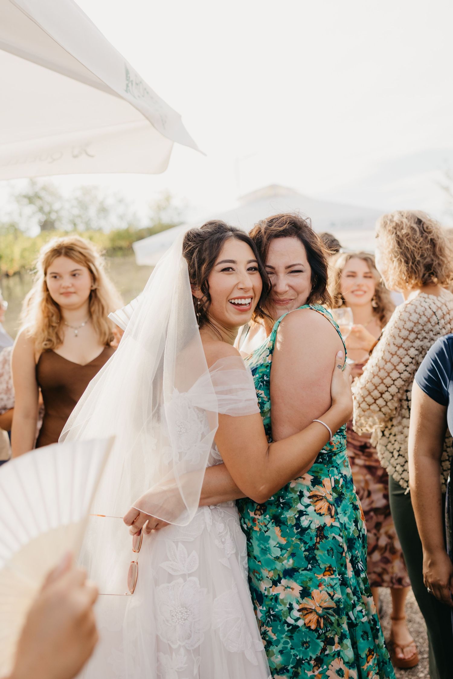 Bridesmaids in turquoise floral dresses embrace the bride in a white wedding gown during an outdoor celebration.