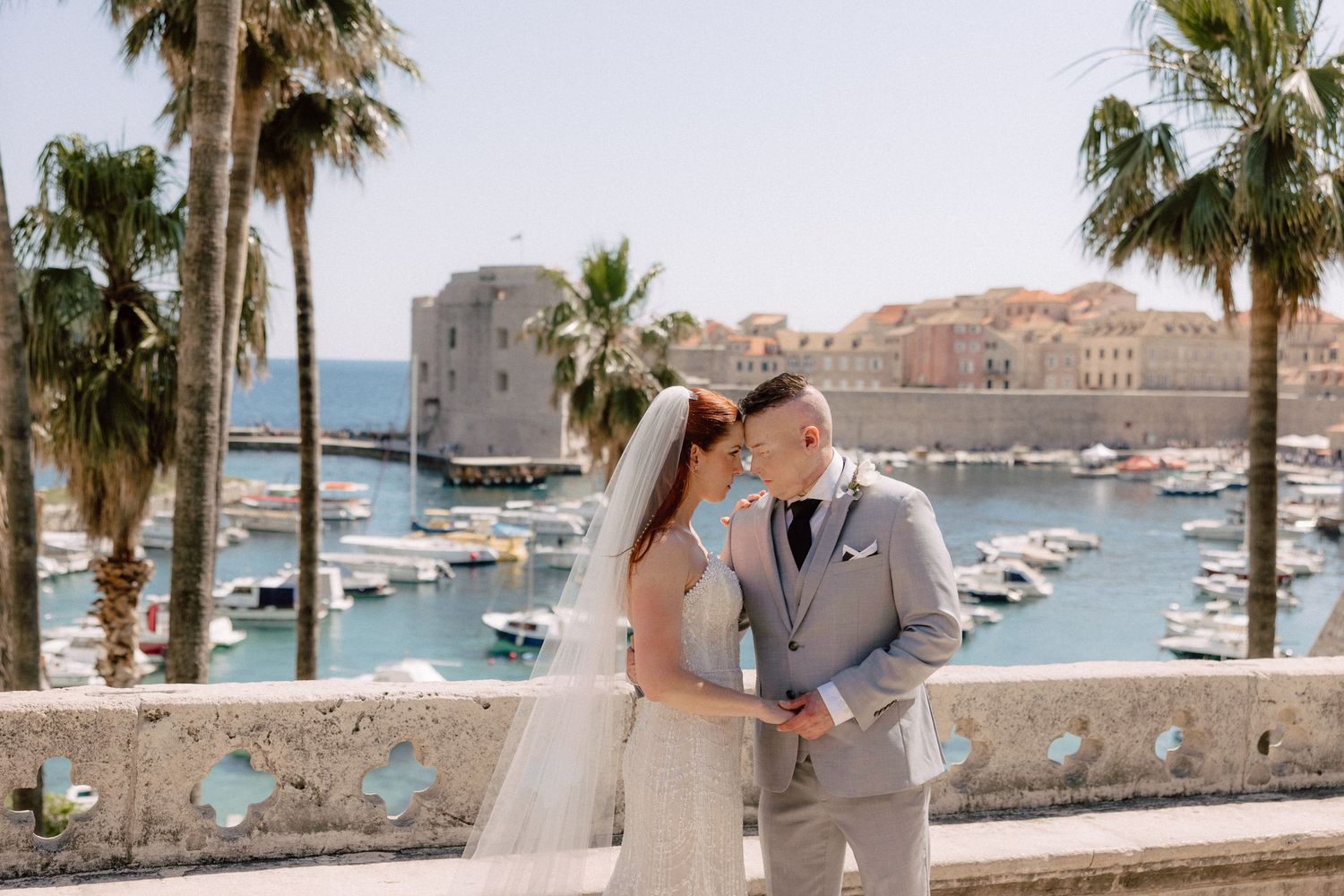 Coastal wedding photography with palm trees and boats in a Mediterranean harbor backdrop.
