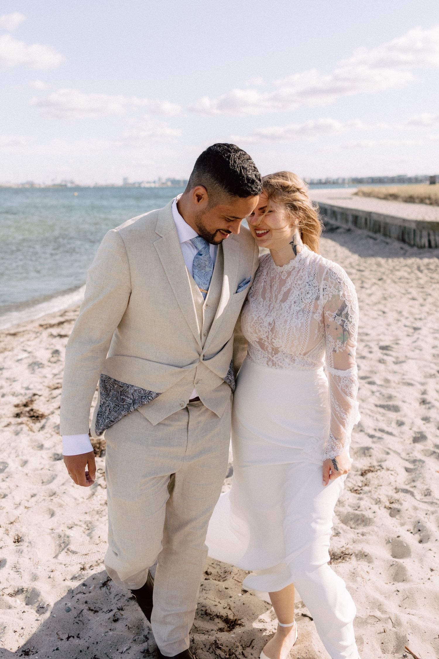 A couple in cream-colored wedding attire embraces on a beach with ocean views.