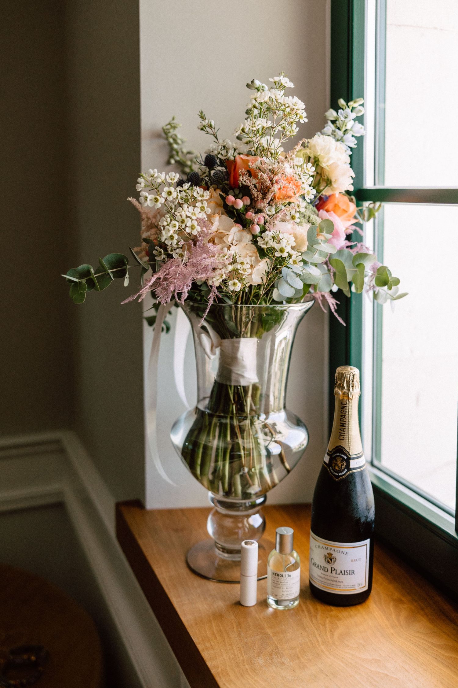 Elegant wedding flat lay with a floral arrangement in a glass vase, champagne bottle, and perfume on a wooden windowsill.