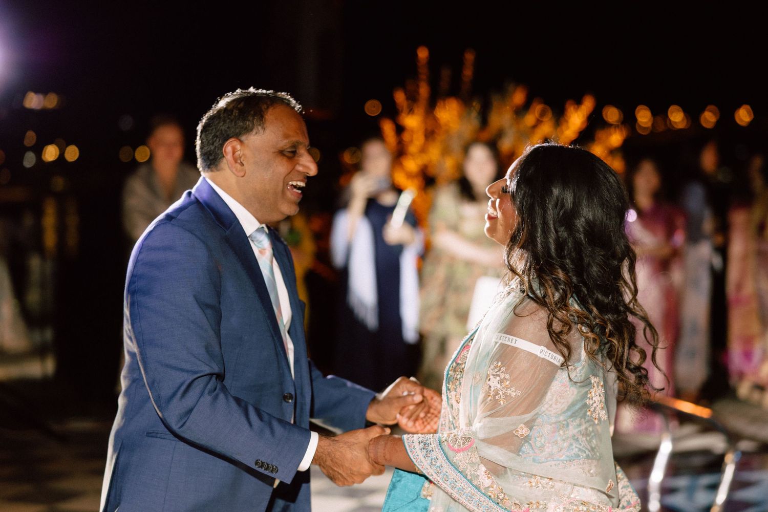 Couple in elegant attire holding hands and smiling at an outdoor evening wedding celebration.