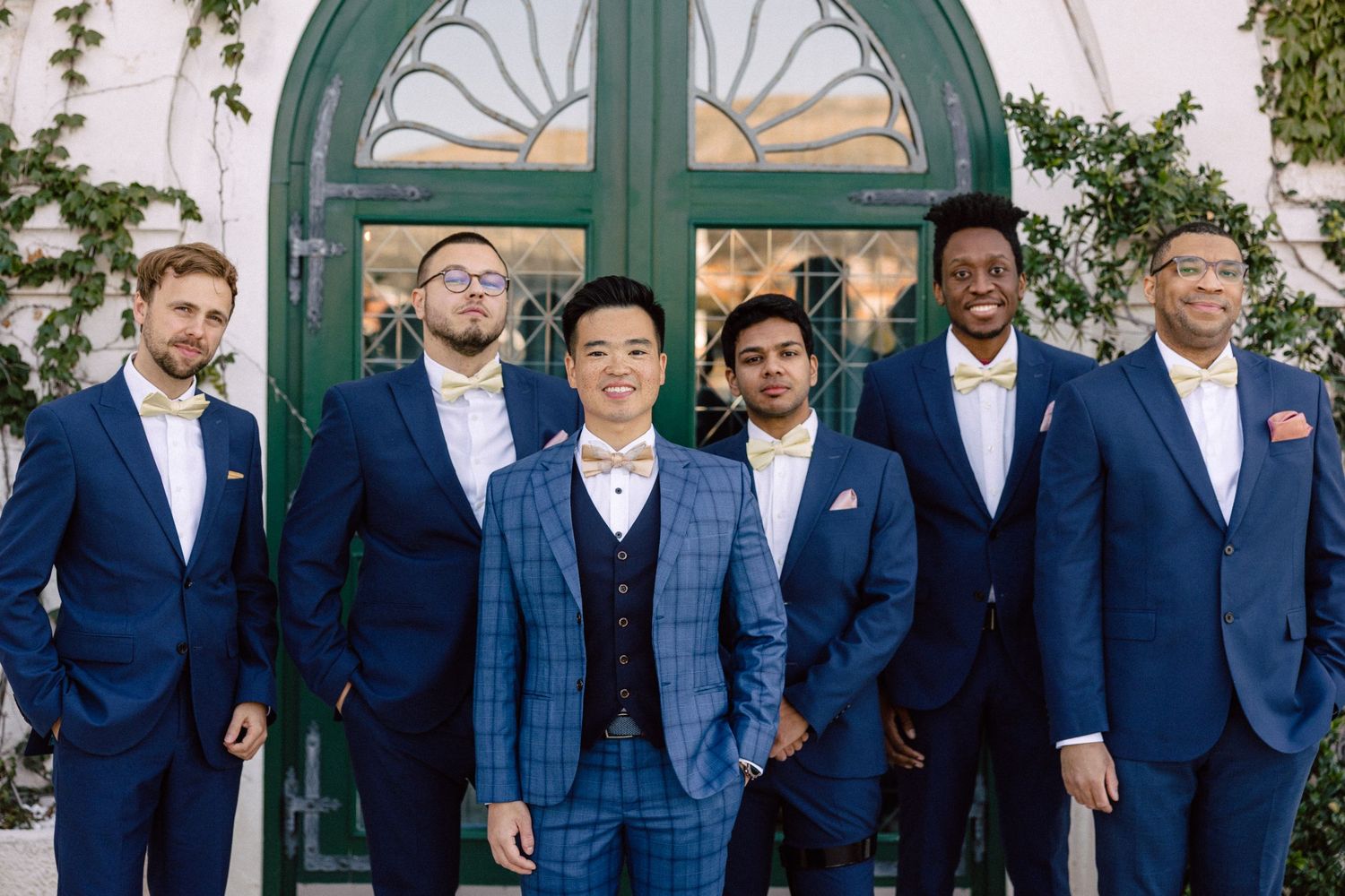 Six groomsmen in navy blue suits with bow ties posing in front of a green ivy-covered door.