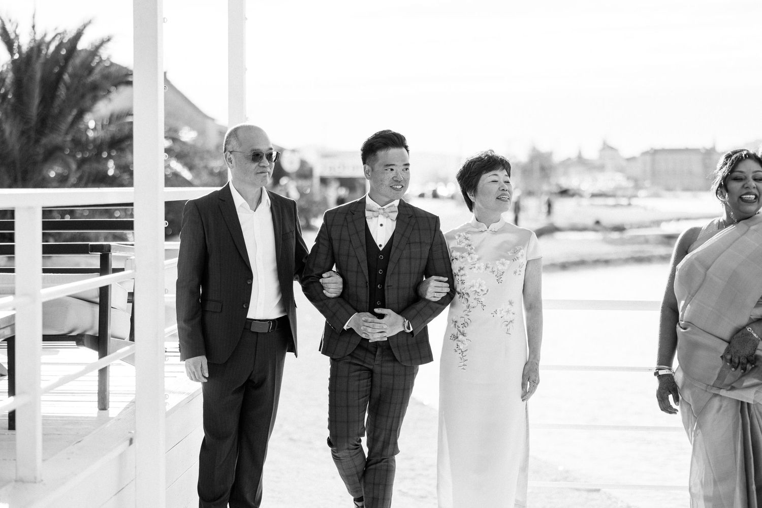 Black and white photo of a groom walking down the aisle escorted by his parents at a seaside wedding.