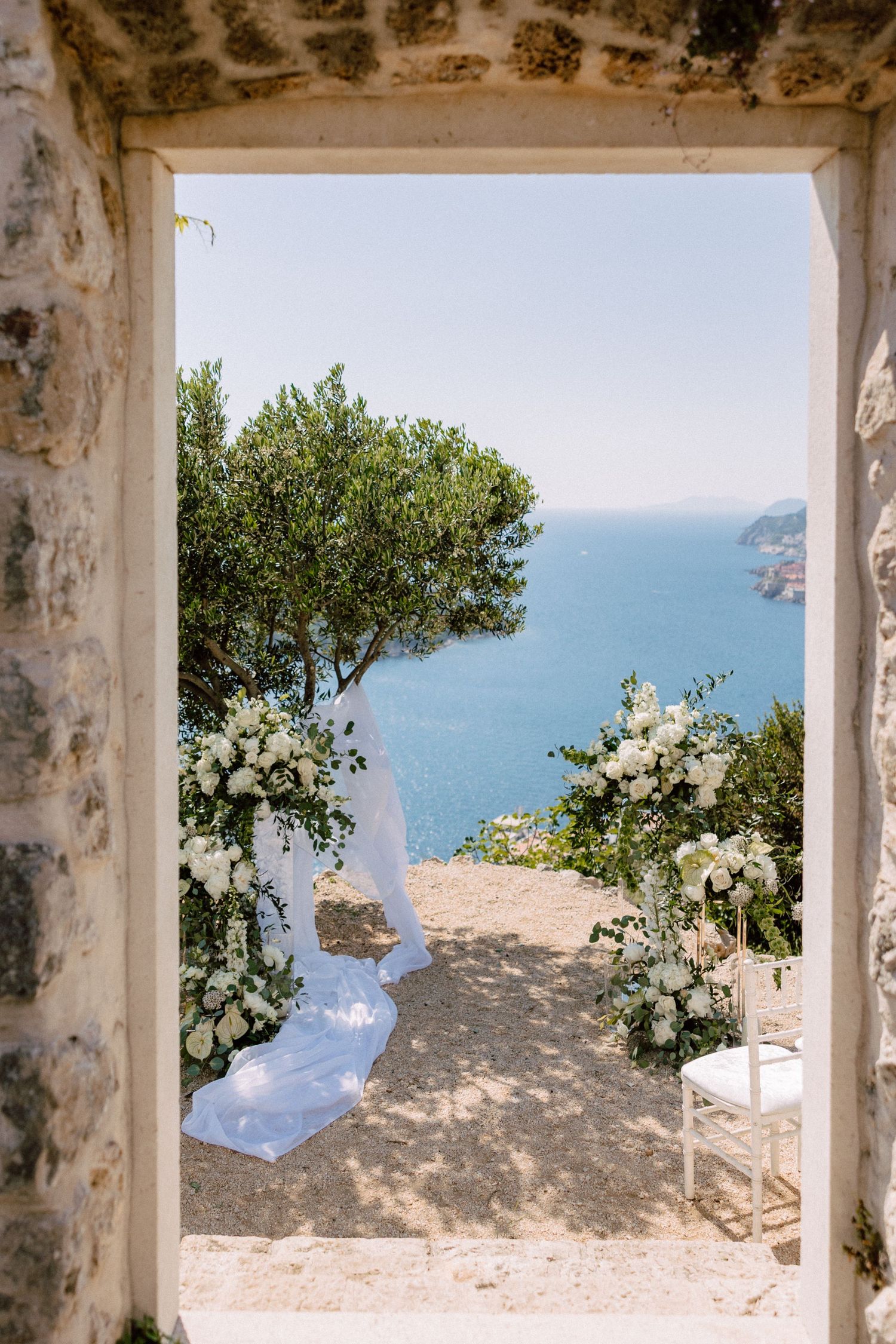 White floral arrangements frame stone archways overlooking the blue Mediterranean Sea.