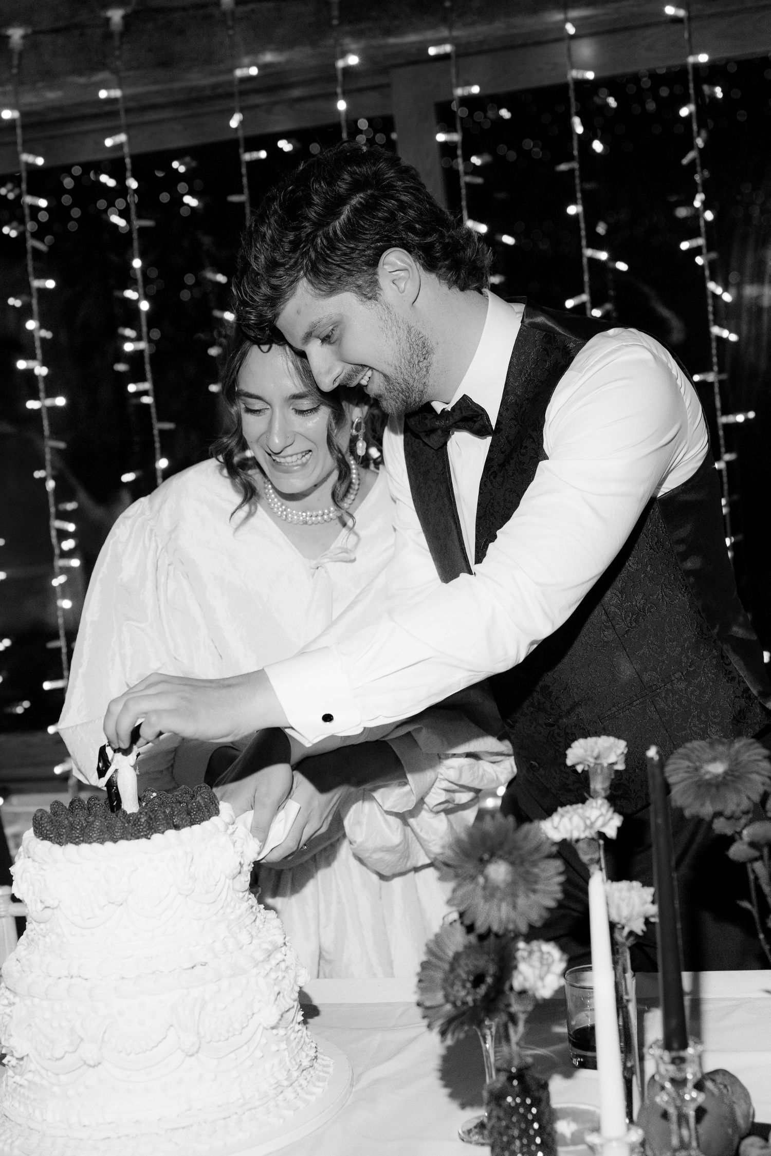 Wedding couple cutting their white tiered cake under string lights.