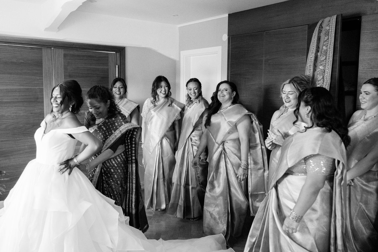 Black and white photo of bride in white gown with bridesmaids in silk sarees laughing together before Indian wedding ceremony.