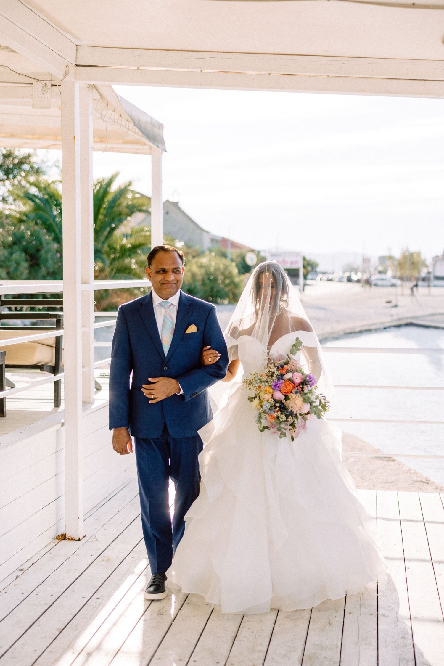 Father walking bride down the aisle in white ball gown with colorful bouquet at waterfront venue.