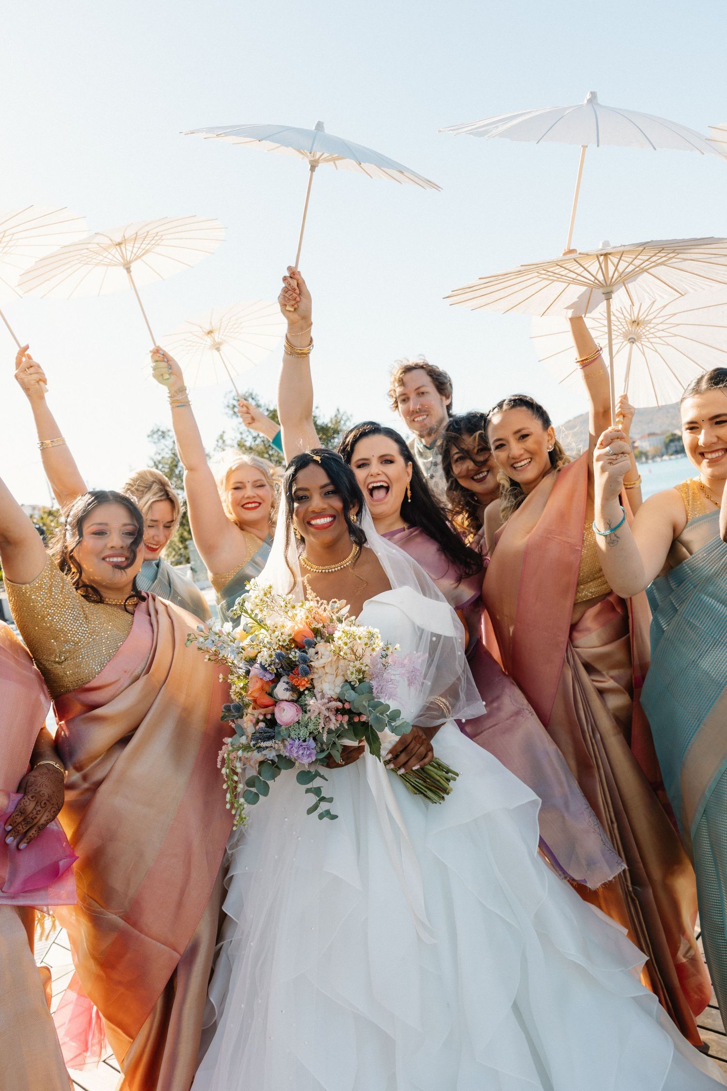Joyful bride with colorful bouquet surrounded by bridesmaids in pink and gold saris holding white parasols.