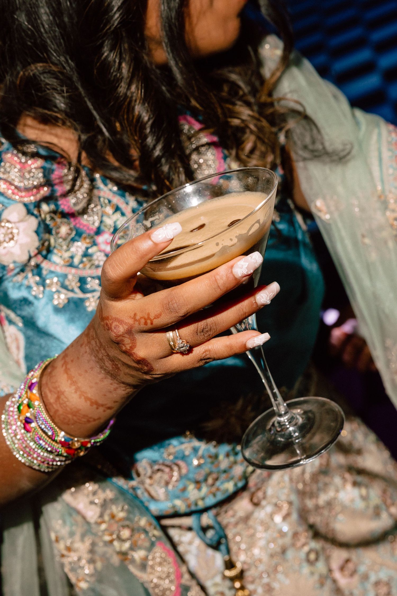 Close-up of henna-adorned hand with bangles holding an espresso martini at a wedding celebration.